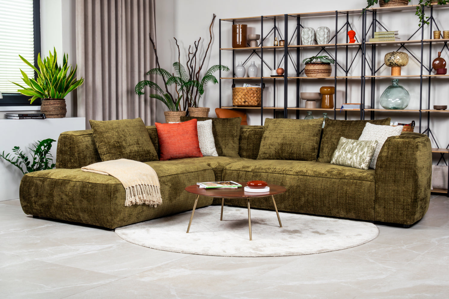 Alyvuogių žalios spalvos kampinė sofa svetainėje prie lentynos